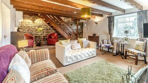 A cozy living room with a stone wall, wooden beams, a fireplace, and a comfortable seating area with a sofa and chairs.