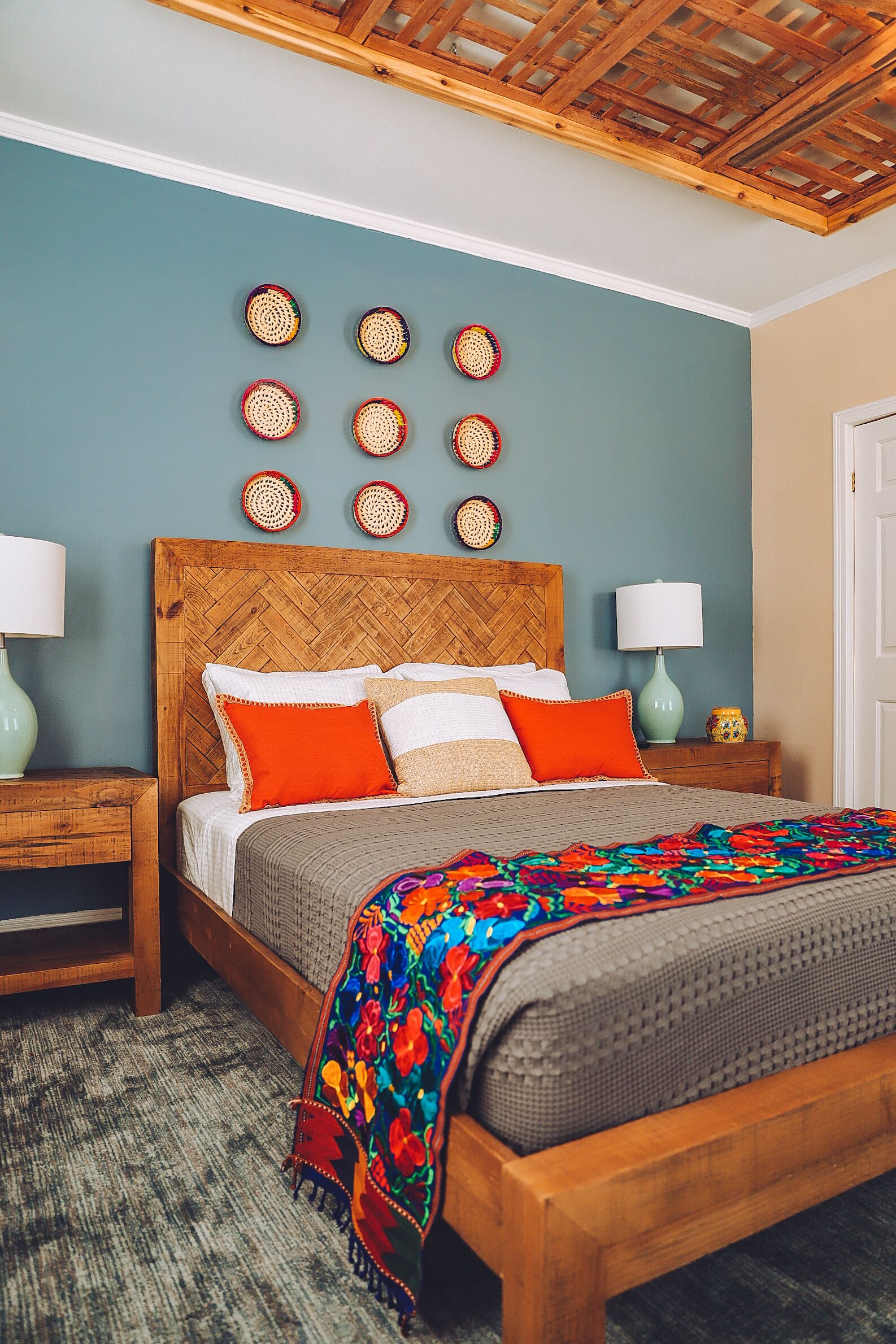 Ein Schlafzimmer mit einem Holz-Kopfteil, einem Bett mit orangefarbenen und weißen Kissen, einer farbenfrohen Decke und einer Wand mit gewebten Kunstwerken.