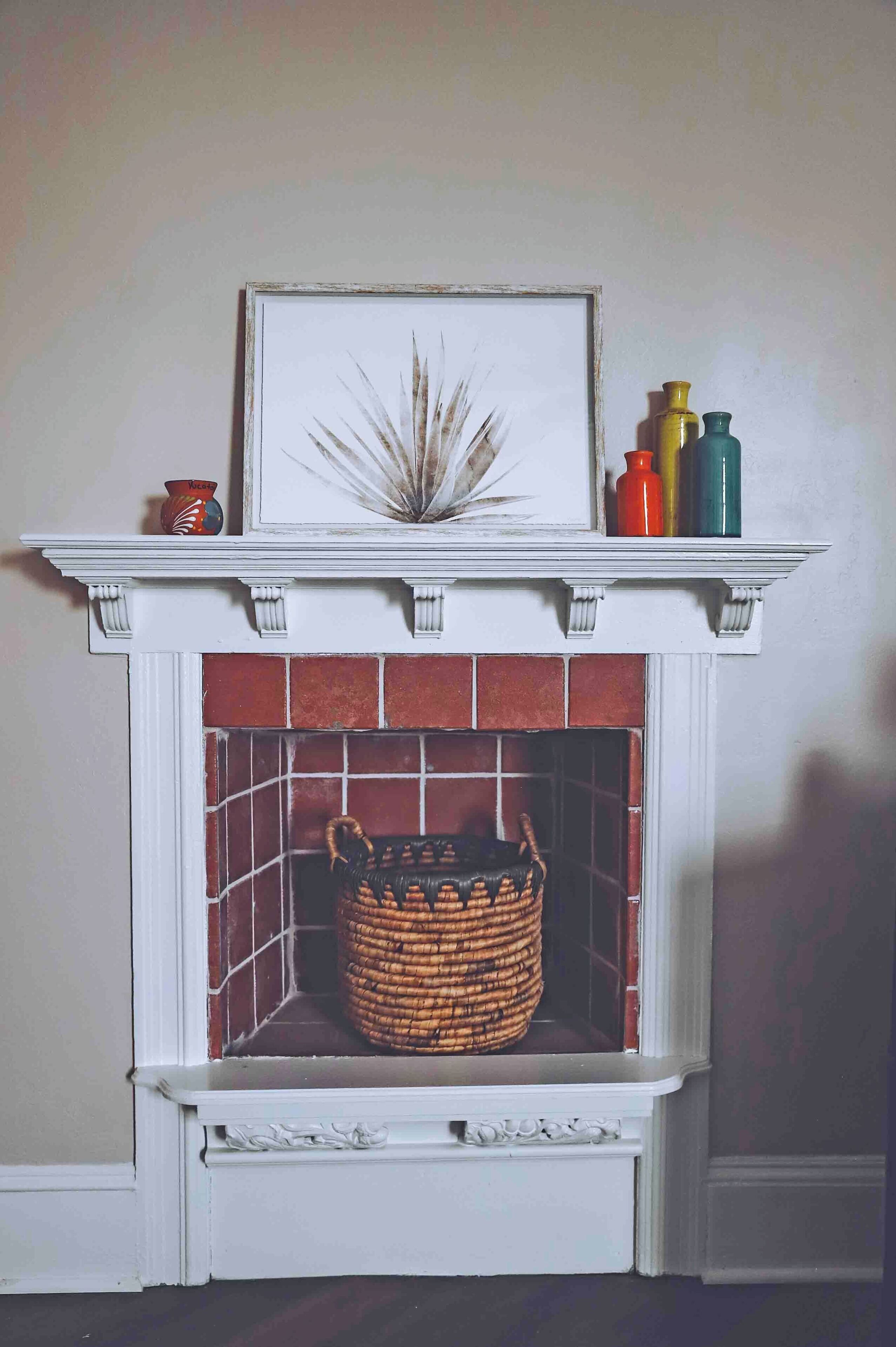 A fireplace with a white mantel, a framed artwork, and a woven basket below.
