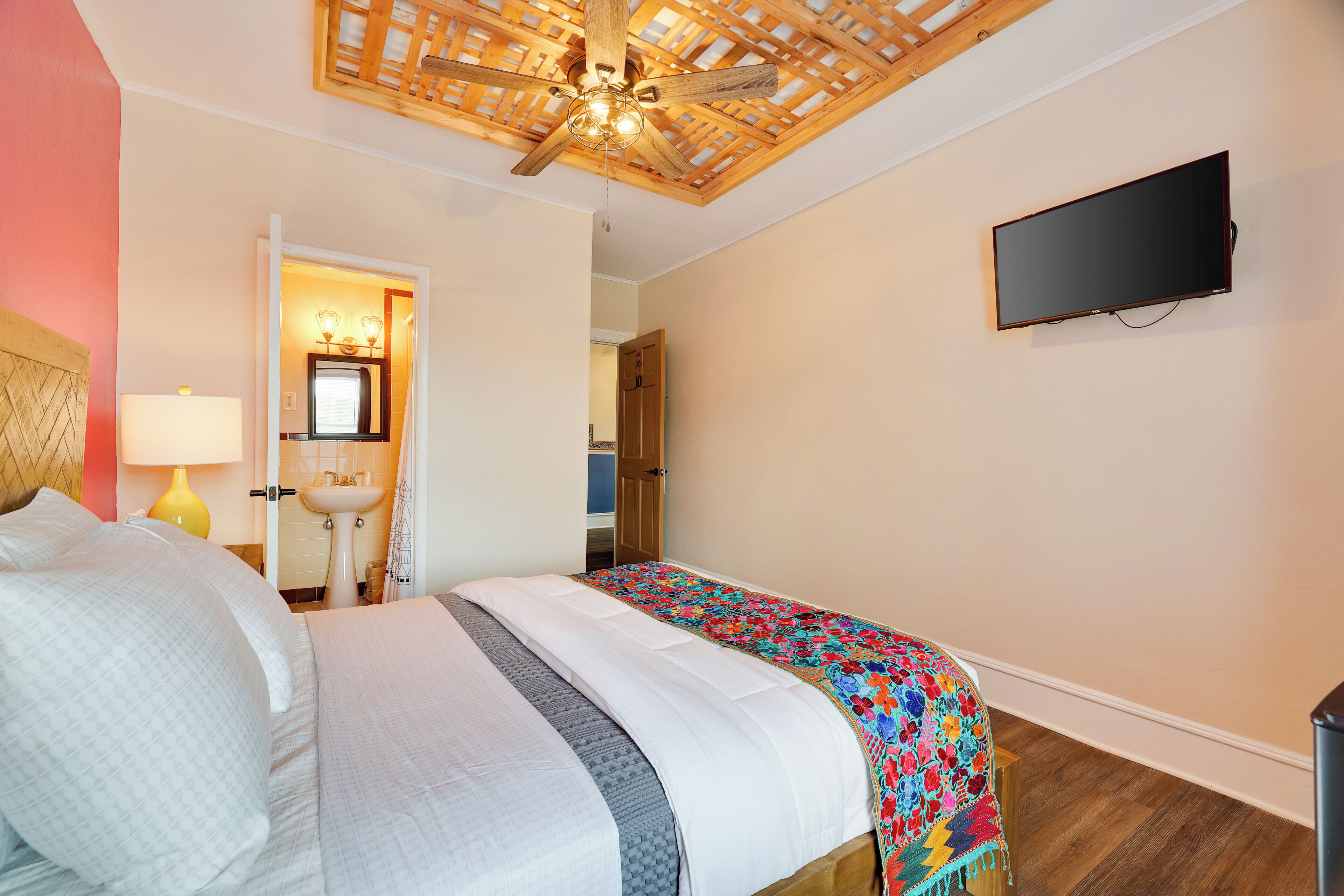 Un dormitorio con una cama, ventilador de techo, televisor y un baño visible a través de una puerta abierta.