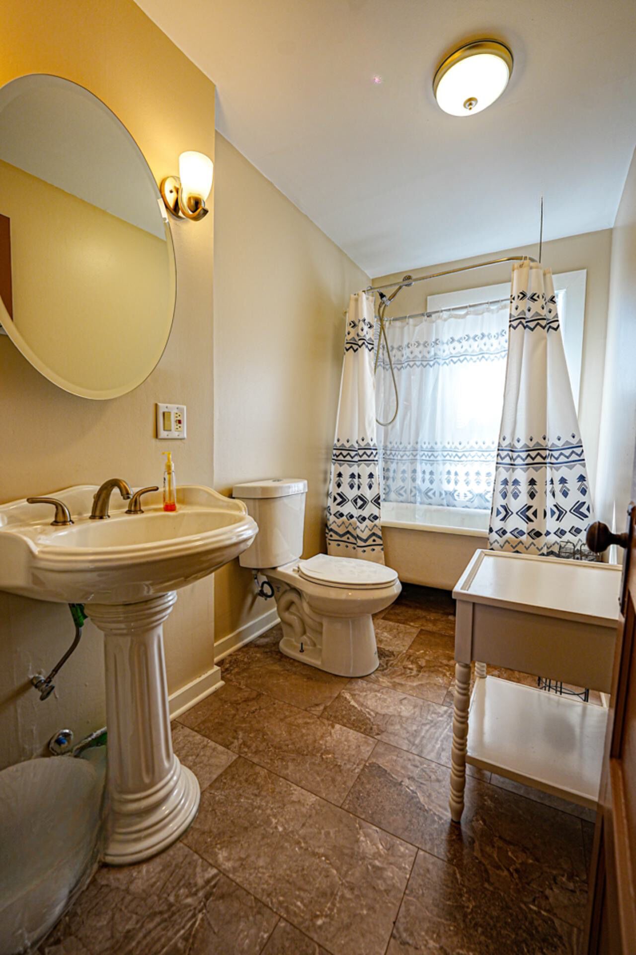 Un baño con lavamanos pedestal, inodoro, bañera con cortina de ducha y ventana con cortinas decoradas.