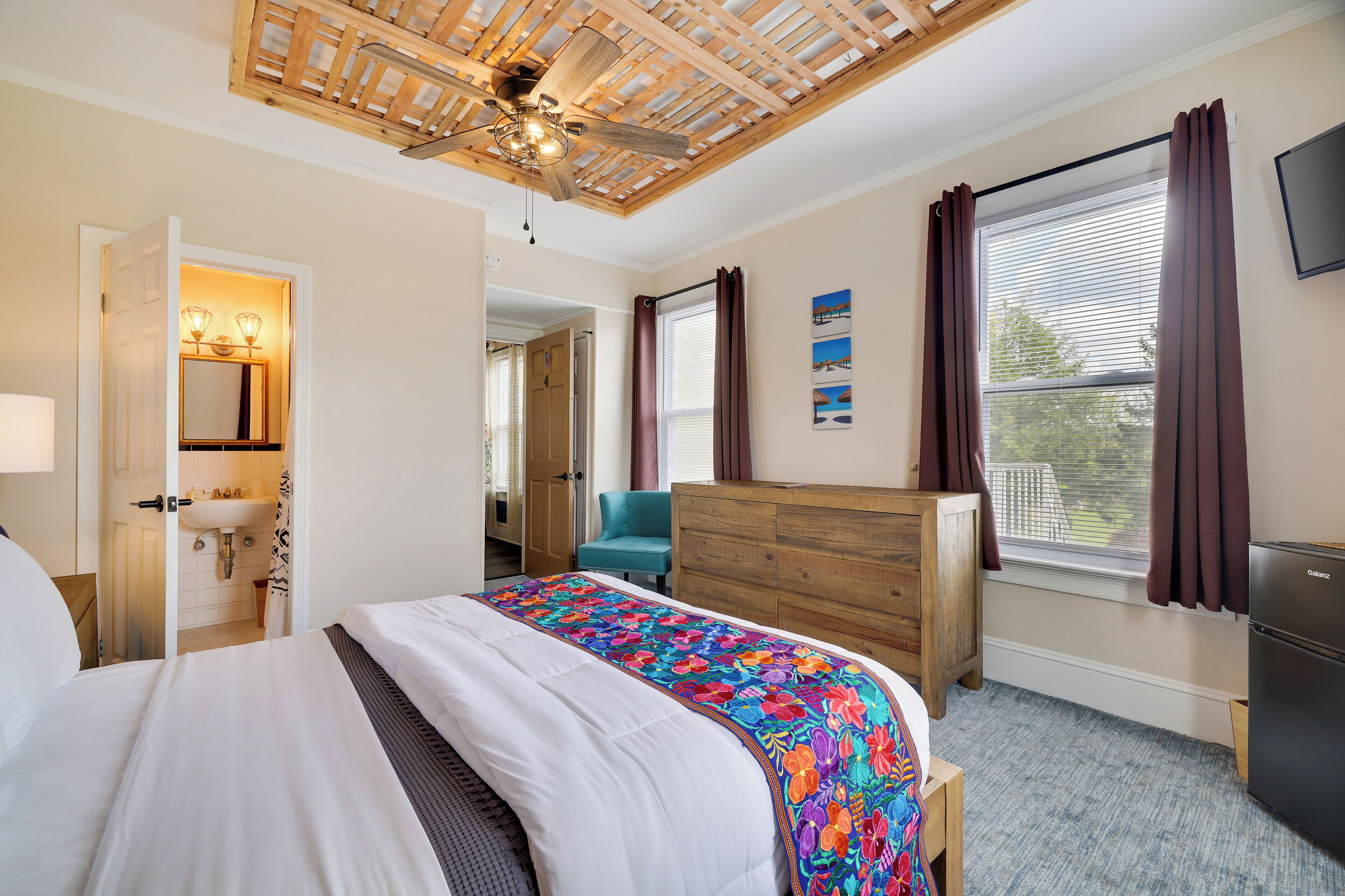 Un dormitorio con cama, cómoda de madera, ventilador de techo, ventana con cortinas y televisor en la pared.