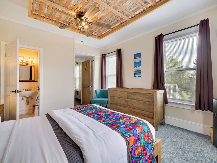Un dormitorio con cama, cómoda de madera, ventilador de techo, ventana con cortinas y televisor en la pared.