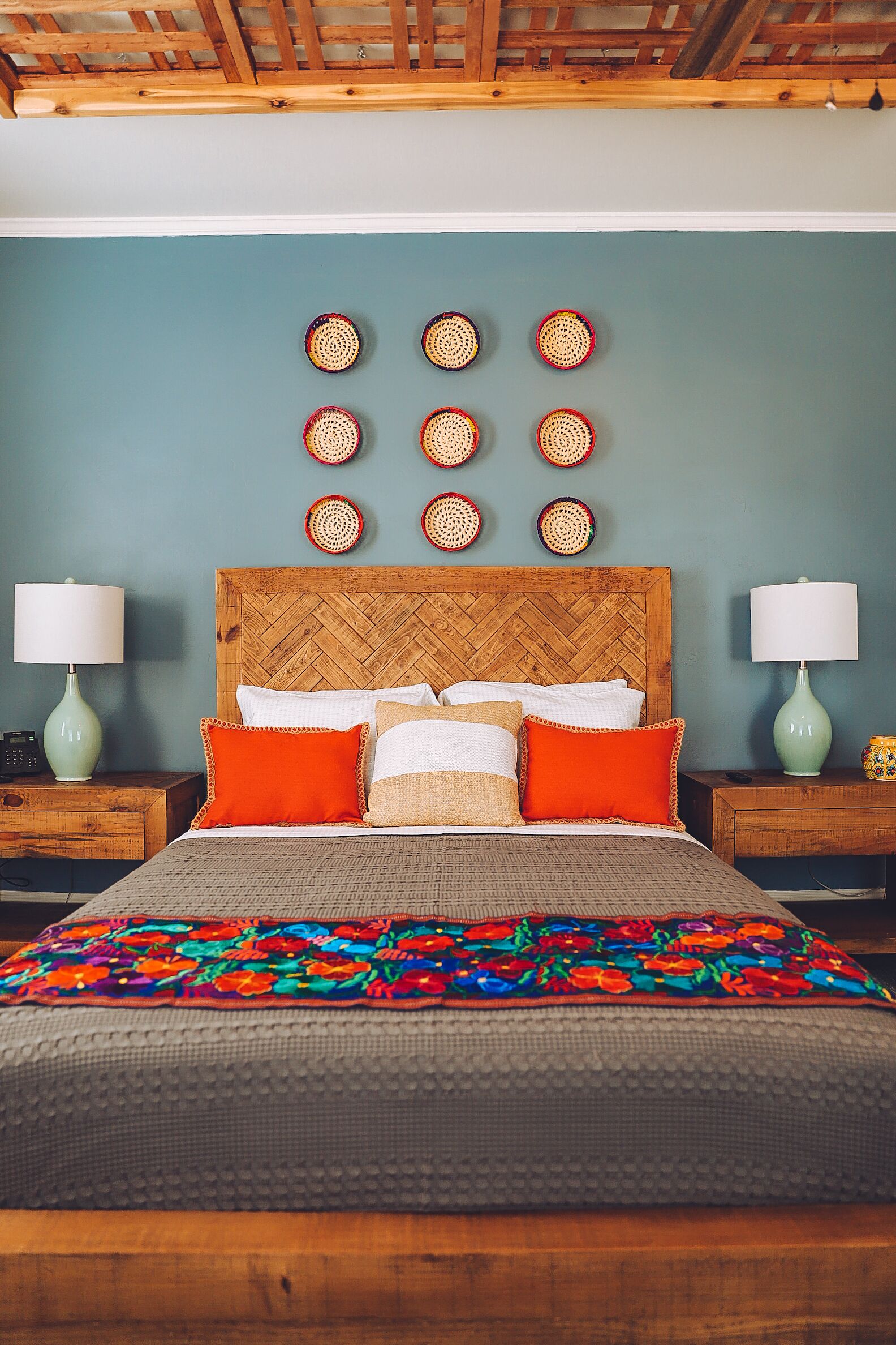 A bedroom with a wooden headboard, a bed with multiple pillows, two bedside tables with lamps, and a wall with circular woven decorations.