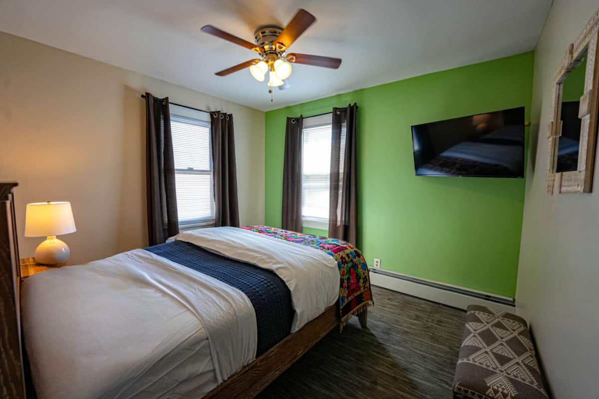 Un dormitorio con cama, ventilador de techo, televisión montada en la pared y ventana con cortinas.