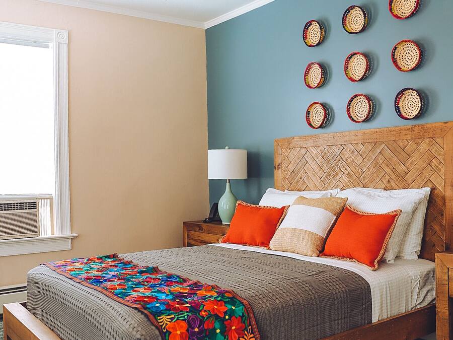 A bedroom with a wooden bed, a colorful blanket, a herringbone-patterned headboard, and woven wall art.