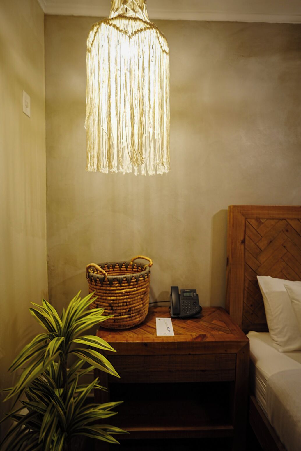 Une chambre avec une table de chevet en bois, un panier en osier, un téléphone sur la table de chevet, une plante et un luminaire suspendu.