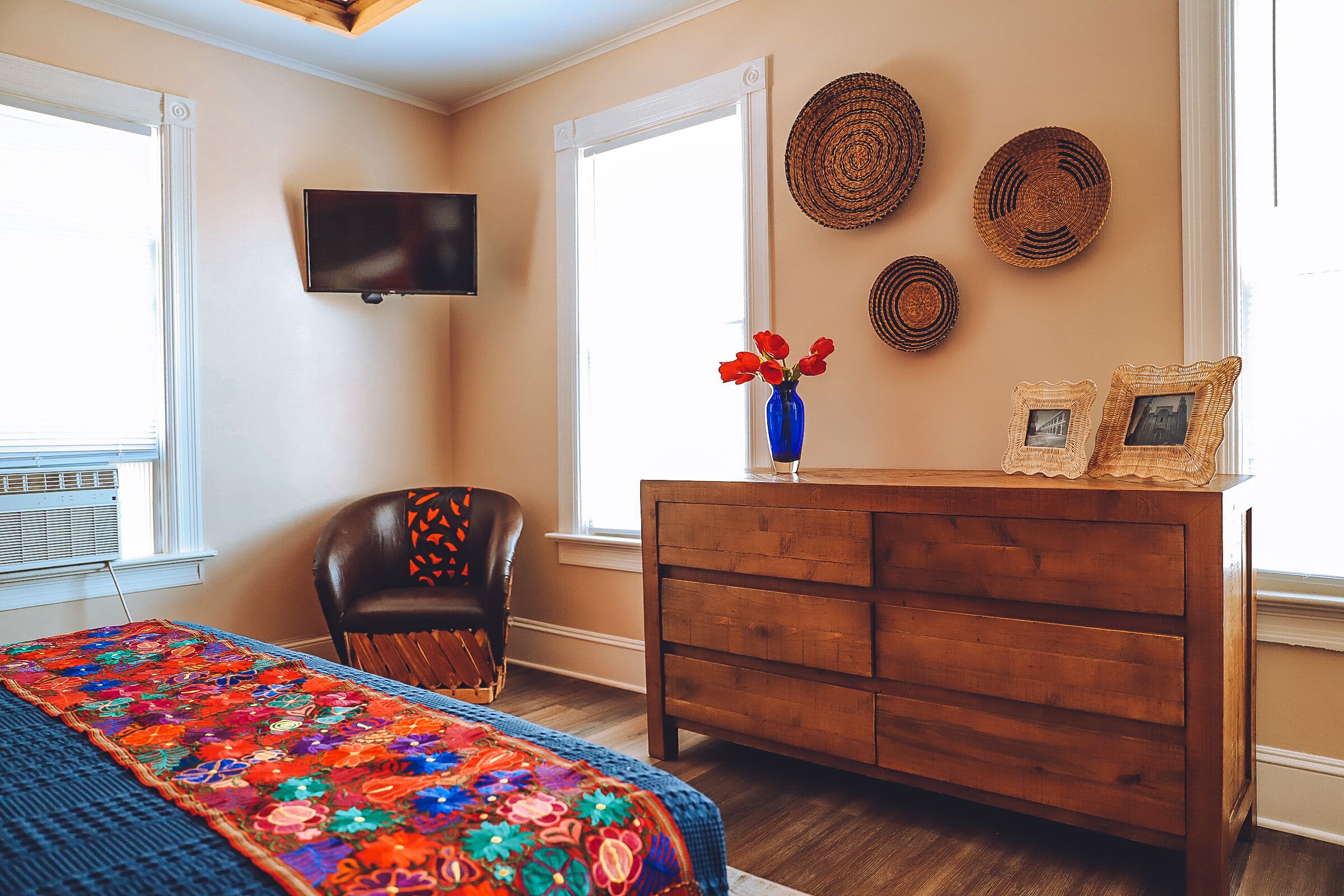 Un dormitorio con un aparador de madera, una silla, un televisor montado en la pared y una ventana con persianas.