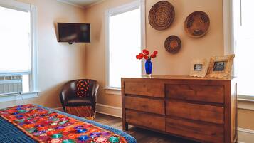 A bedroom with a wooden dresser, a chair, a TV mounted on the wall, and a window with blinds.