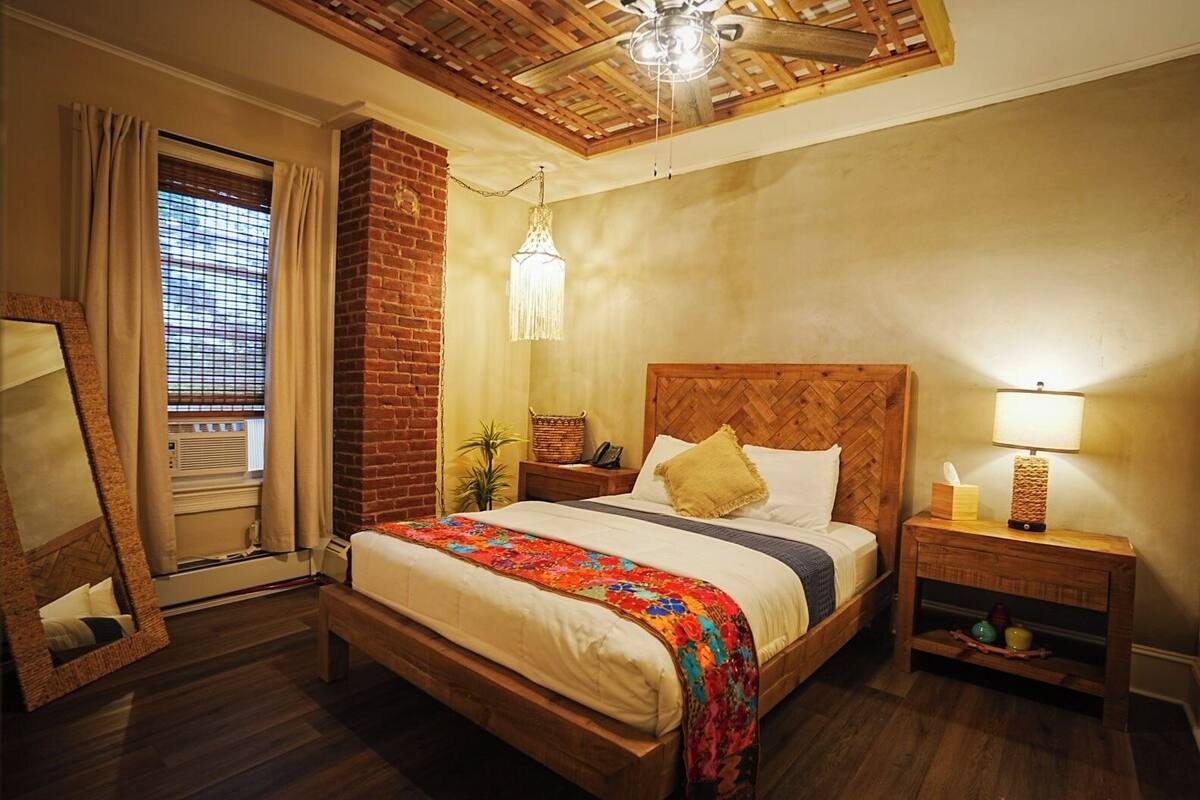 Un dormitorio con cama de madera, pared de ladrillo, ventilador de techo, lámpara colgante, mesita de noche con lámpara y ventana con cortinas.
