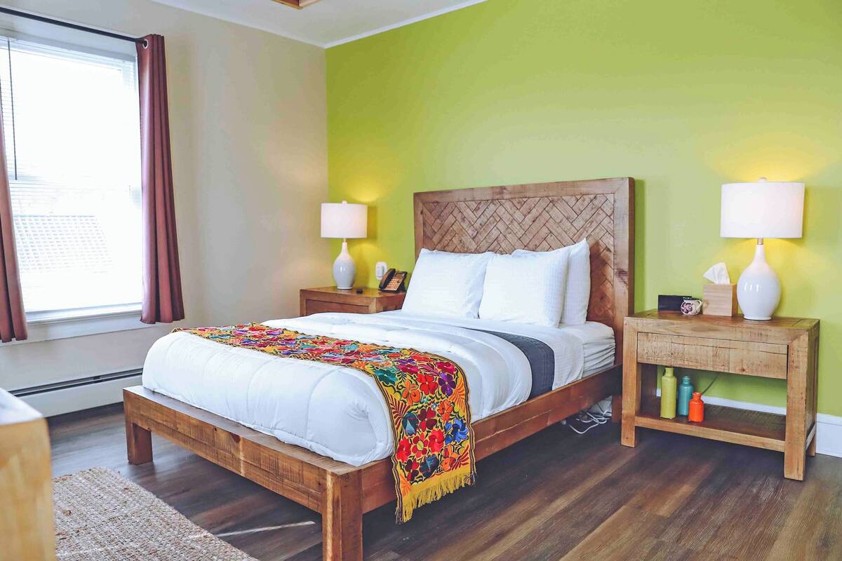 Una cama con estructura de madera, ropa de cama blanca y una colcha colorida. Hay dos mesitas de noche con lámparas y un mueble con un teléfono y una caja de pañuelos.