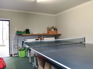 A ping pong table in a room with a door, a green bag, and a shelf with various items.