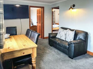 Een kamer met een houten eettafel, twee zwarte leren stoelen, een zwarte leren bank met gestreepte kussens, een flatscreen televisie aan de muur en een wandlamp.