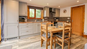 A modern kitchen with grey cabinets, a wooden dining table with chairs, and a window providing natural light. - Little Lodge (King's Lynn)