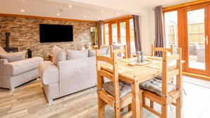 A living room with a stone wall, a flat-screen TV, a sofa, and a wooden dining table with chairs. - Little Lodge (King's Lynn)