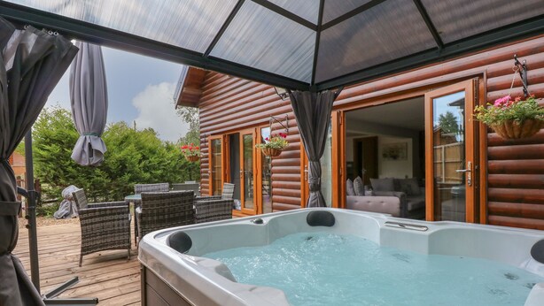 A hot tub in a covered outdoor area with wooden deck and wicker furniture. - Little Lodge (King's Lynn)