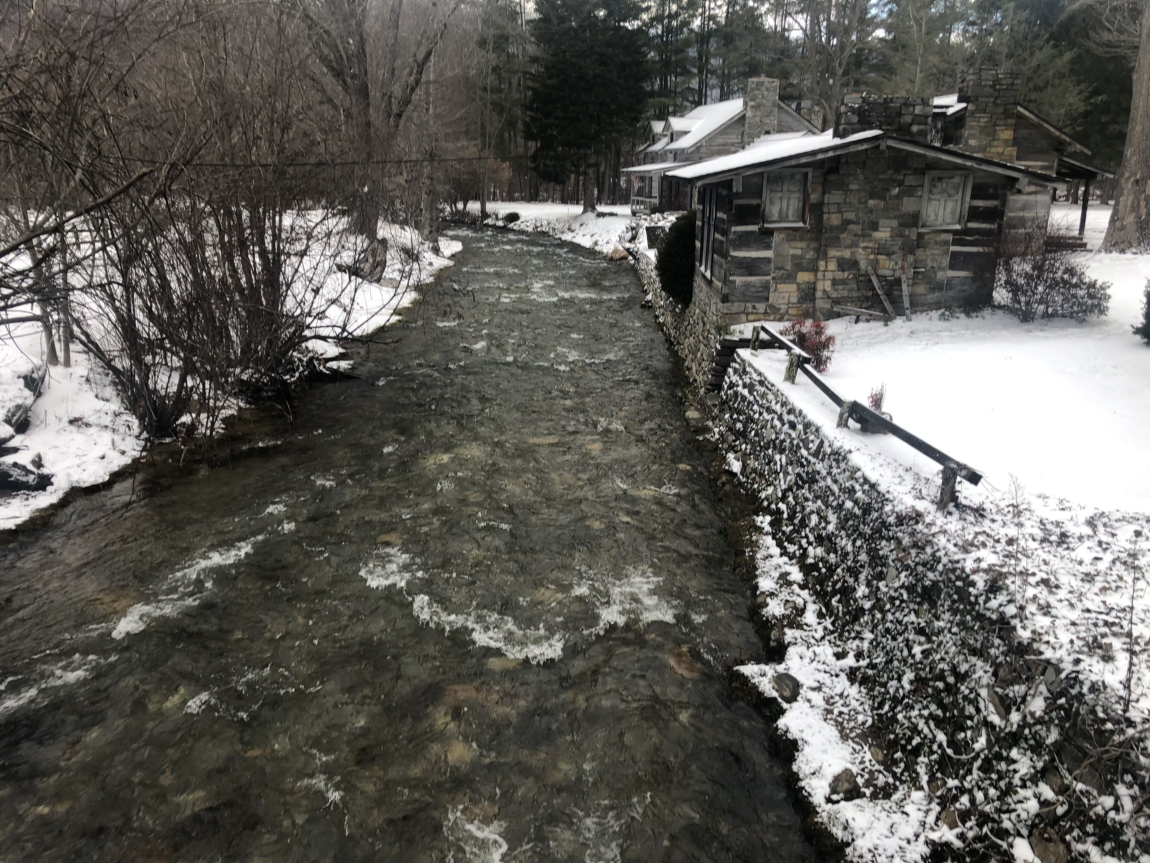 Un pont de pierre enjambe un ruisseau enneigé dans une zone boisée, avec un bâtiment en pierre sur la droite.