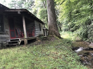 Un chalet rustique doté d’un porche, entouré d’une forêt dense et traversé par un petit ruisseau.
