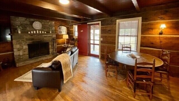 A cozy log cabin interior with a stone fireplace, wooden dining table, and a dog resting on a chair.