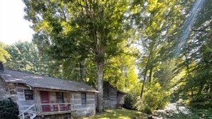 A rustic cabin with a porch, surrounded by dense forest and a small stream.