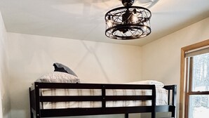 A bunk bed with a checkered bedspread and pillows, a wooden nightstand with a lamp, and a window with a view of trees. - Lost on the Lake-Cozy Cottage with a Lakeview (Wetmore)