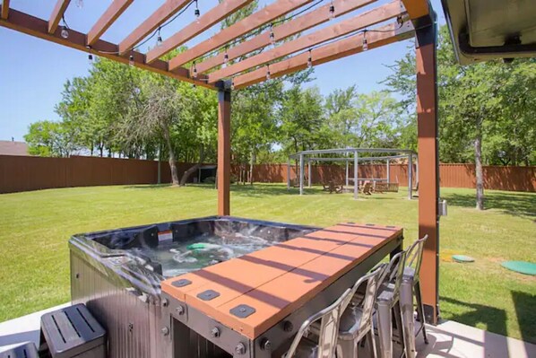 A hot tub under a wooden pergola with a grassy yard and trees in the background. - The Rooftop (Denison)