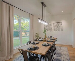 A dining area with a long wooden table set for six, surrounded by chairs. There are two wall-mounted light fixtures above the table and a large window with curtains on the left.