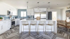 Une cuisine moderne avec des armoires blanches, un grand îlot central doté de tabourets, des appareils en acier inoxydable et un parquet en bois disposé en chevrons.