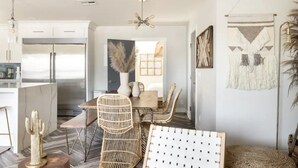 Une cuisine moderne avec des meubles blancs, un réfrigérateur en acier inoxydable et un coin repas comprenant des chaises en osier et une table en bois.