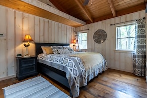 A wooden-paneled bedroom with a bed, two bedside lamps, a round wall decoration, and a window with curtains. - Sweet Sapphire Getaway! Enjoy the mountains!! (Sapphire)