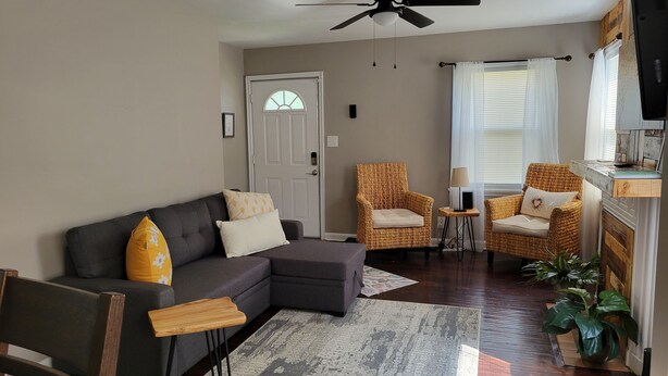 A living room with a dark grey sectional sofa, two wicker armchairs, a small wooden side table, a ceiling fan, and a large window with white curtains. - Clean, quiet and perfectly located in the city.  Near downtown. (Columbus)