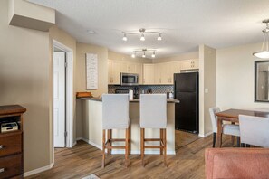 A modern kitchen with wooden cabinets, a black refrigerator, and a dining area with two bar stools. - Hart of it All | Top Floor | Pool | Hot Tub | Gym (Invermere)