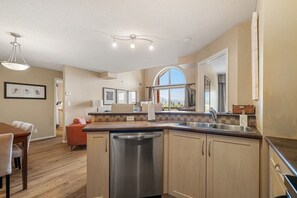 A modern kitchen with a dishwasher, a sink, and a dining area with a table and chairs. - Hart of it All | Top Floor | Pool | Hot Tub | Gym (Invermere)