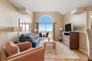 A spacious living room with a large arched window, a blue sofa, a wooden coffee table, and a television on a wooden cabinet. - Hart of it All | Top Floor | Pool | Hot Tub | Gym (Invermere)