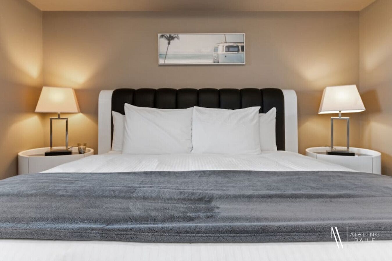A neatly made bed with a dark gray blanket, two bedside tables with lamps, a headboard, and a framed picture on the wall. - Lake Life Livin | Lake View | Pool | Hot Tub | Gym (Invermere)