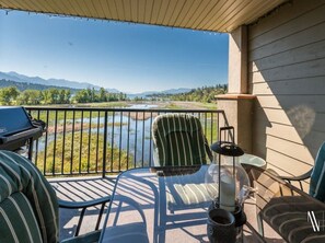 A balcony with a view of a lake and mountains, furnished with striped chairs and a table, and a grill. - Lake Life Livin | Lake View | Pool | Hot Tub | Gym (Invermere)