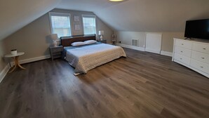 Une chambre avec un plafond en pente, un lit recouvert d’une couvre-lit à motifs, des tables de chevet avec des lampes, un téléviseur posé sur une commode et une fenêtre avec des stores.