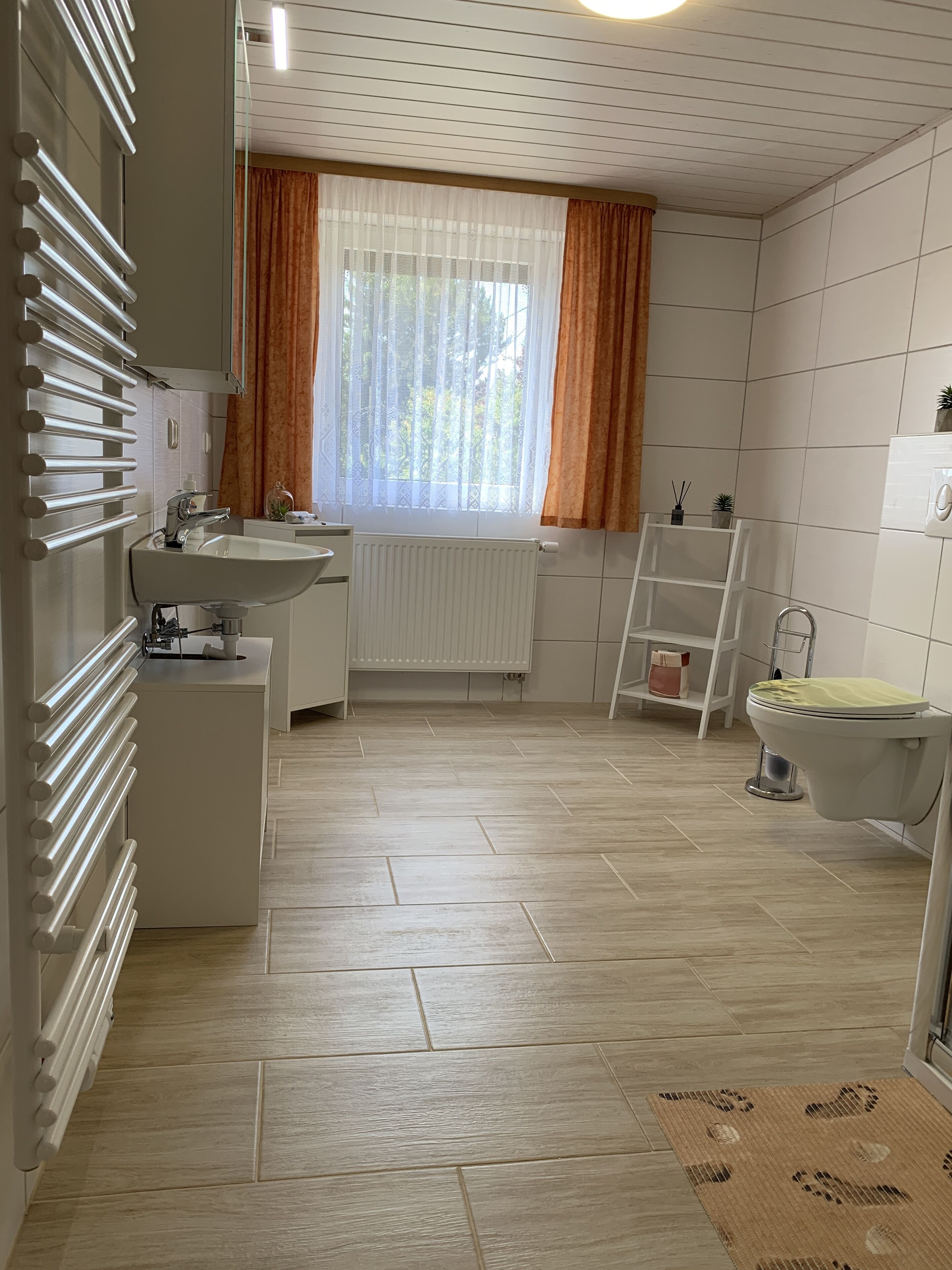 Ein Badezimmer mit weißem Waschbecken, Toilette und Heizkörper. Es gibt ein Fenster mit Vorhängen, einen Spiegel und ein Regal mit Pflegeprodukten.