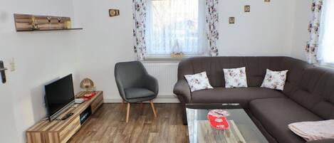 A living room with a sofa, a glass coffee table, a TV on a wooden stand, a chair, and a window with curtains.