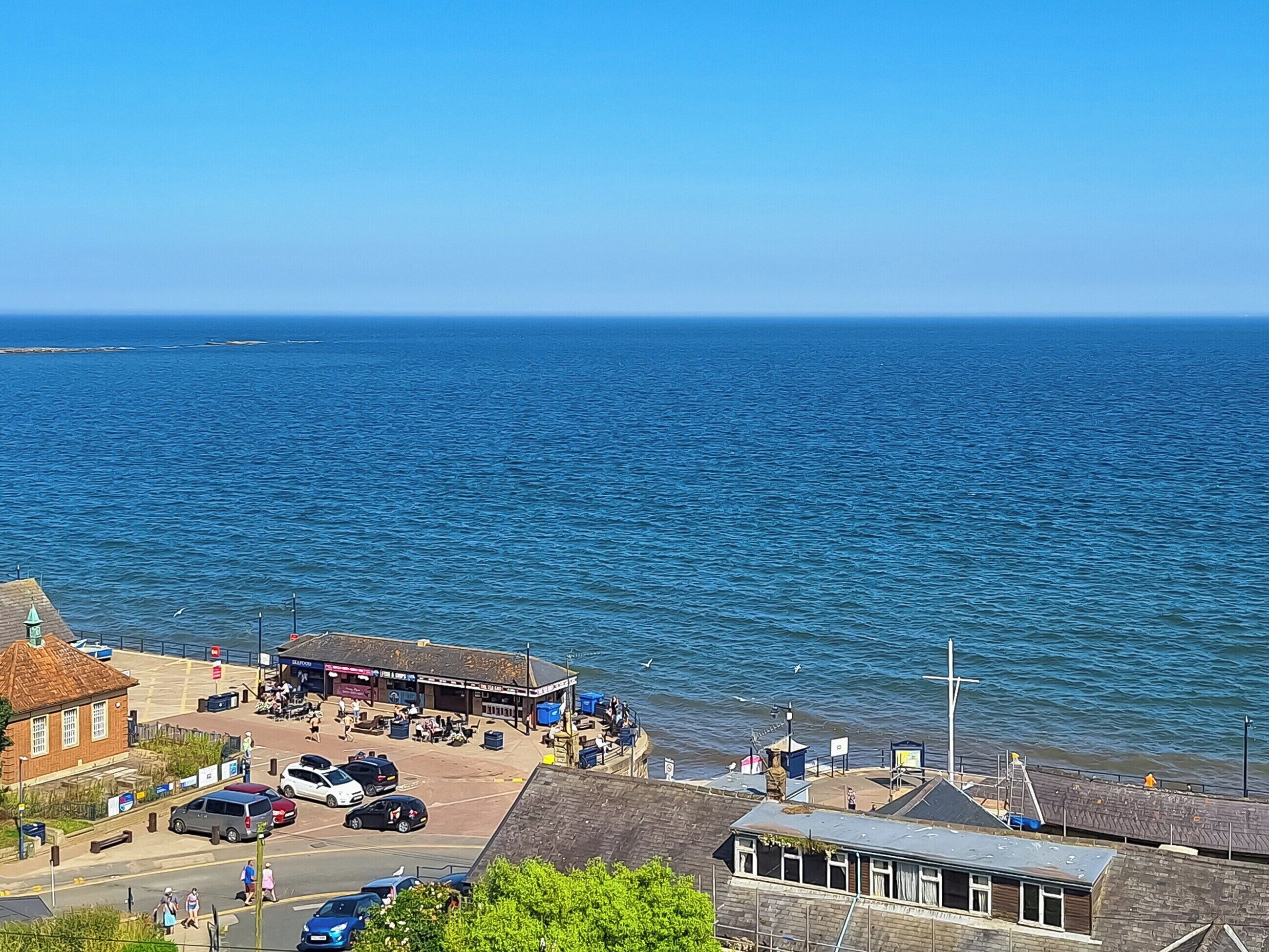 A coastal town with a pier, a small building with a sign, and a road with parked cars.