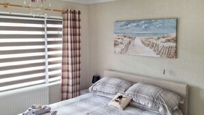 A bedroom with a bed, a nightstand, a window with blinds, a painting on the wall, and a chandelier.