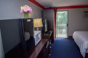 Quarto de hotel com cama, escrivaninha com computador, televisão, micro-ondas e um vaso com flores.