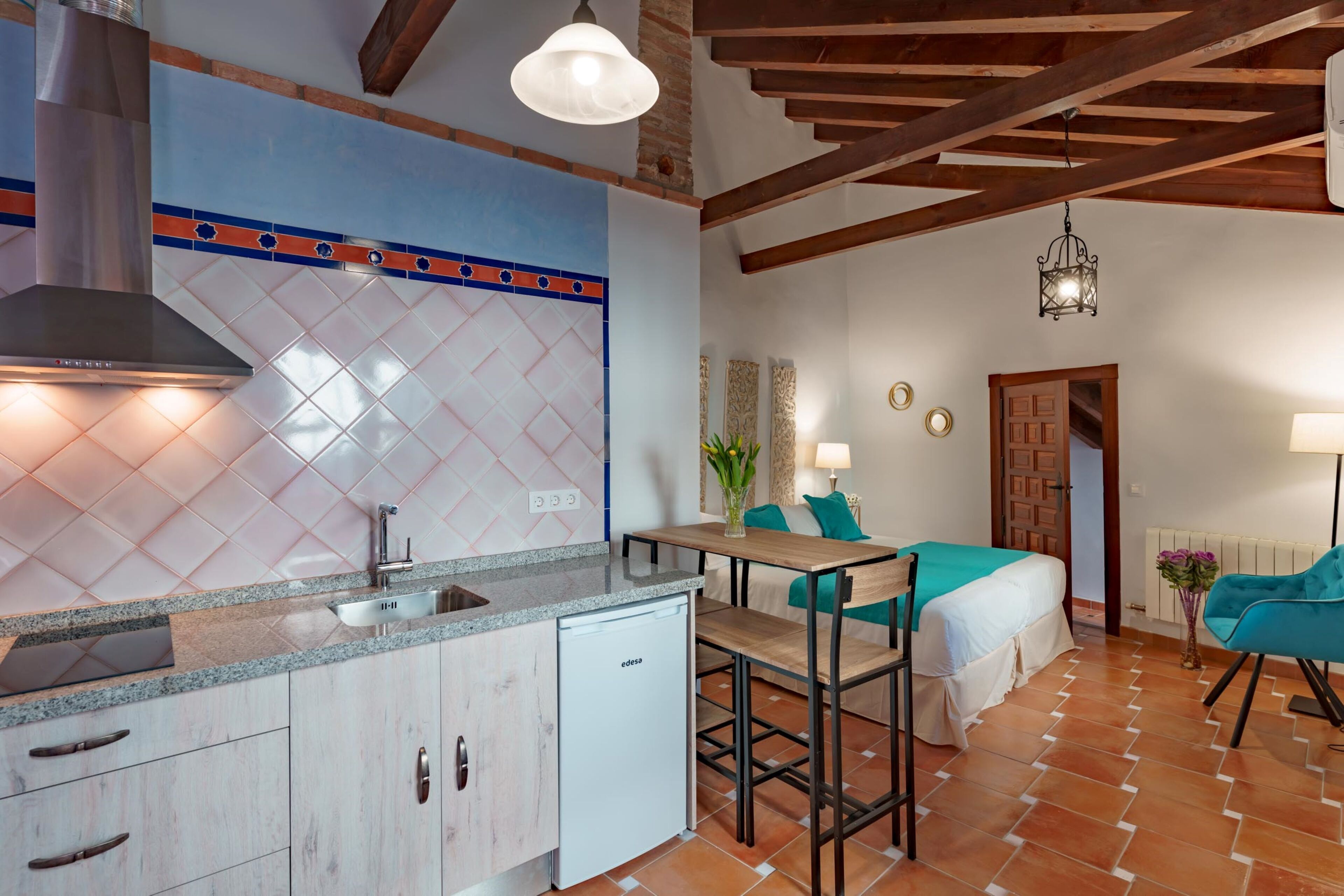 A compact kitchen with a built-in dishwasher, a dining area with a small table and chairs, and a bedroom with a bed and a nightstand.