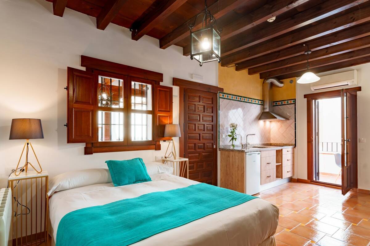 A bedroom with a bed, a nightstand, a lamp, a window with wooden shutters, and a view of a kitchen and balcony.