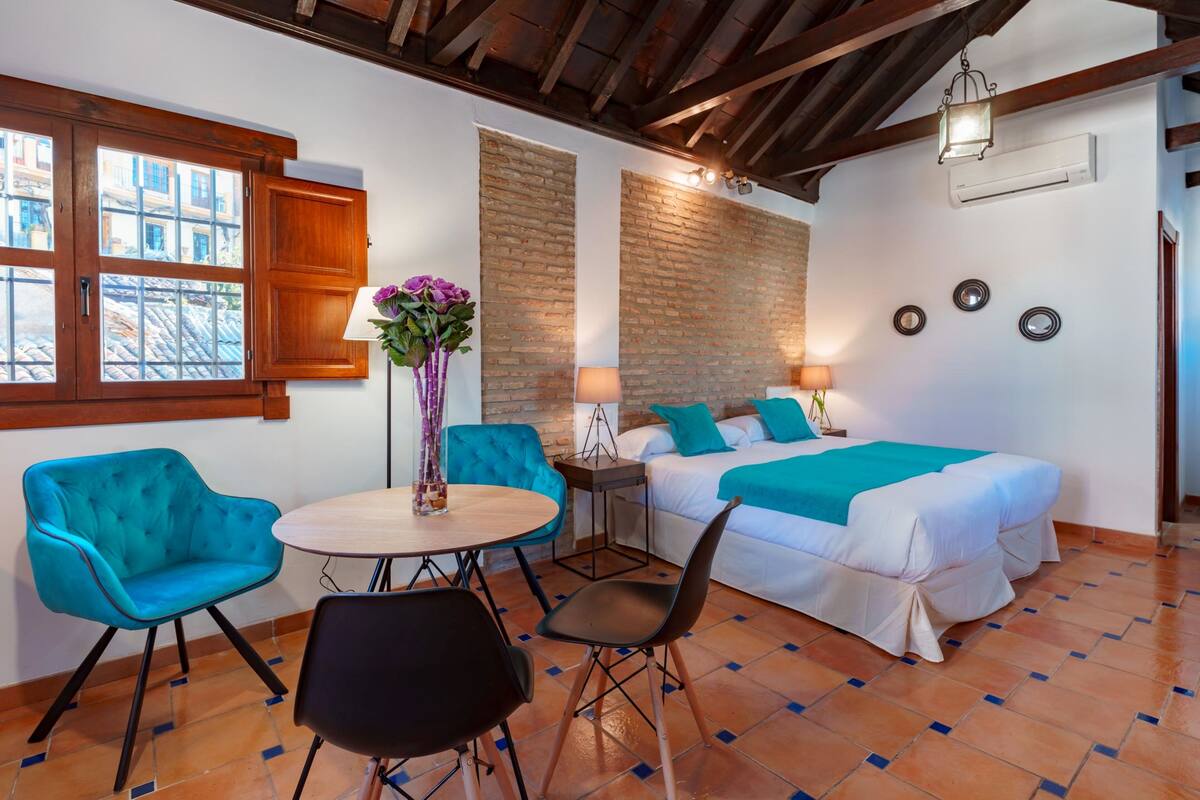 A room with a bed, a table with chairs, a window, and a brick wall.