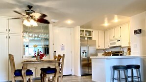 A modern kitchen with white cabinets, a stainless steel refrigerator, a microwave, and a ceiling fan. There is a dining area with a wooden table and chairs. - Pearl on the Strand - Ocean and Village Views at Oceanside Beach! (Oceanside)