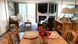 A living room with a dining table set for four, a fireplace, a TV, and a view of the outdoors. - Pearl on the Strand - Ocean and Village Views at Oceanside Beach! (Oceanside)