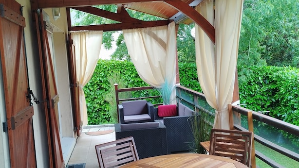 A covered outdoor seating area with a wooden table and chairs, a cushioned sofa, and a view of greenery. - Beautiful view over the heights of Sarlat in a quiet, not overlooked location with air-conditioned swimming pool. (Sarlat-la-Canéda)