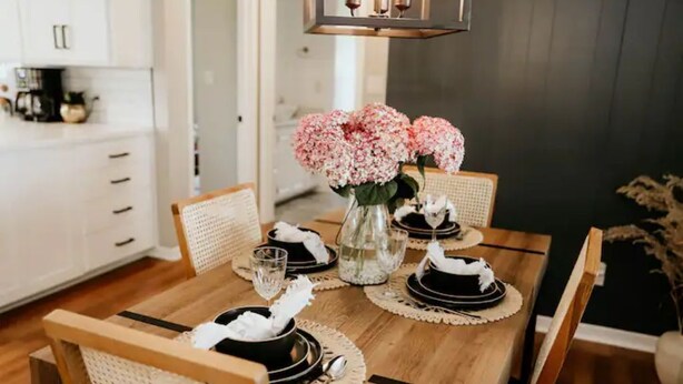 Mesa de comedor preparada para cuatro personas, con sillas blancas, un florero de flores rosas y una lámpara colgante moderna.