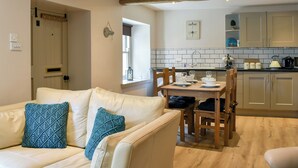 A cozy living room with a beige sofa and two patterned cushions, a wooden dining table with chairs, and a kitchen with wooden cabinets and a tiled backsplash. - 2 bedroom accommodation in Hawes (Hawes)